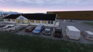 Cars, pickup trucks, and a white van stand outside a low white building, with a warehouse and snow-covered mountains behind.
