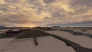 Snow surrounds airport buildings, crowded parking lots, and a curving access road beneath golden clouds and a low sun.
