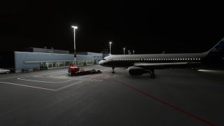 A passenger jet sits beside a floodlit terminal at night, with illuminated cabin windows and a tow tractor near its nose.