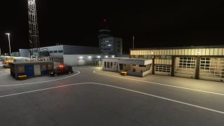 Airport service buildings and a control tower stand illuminated at night beside garage bays and a van with red roof lights.