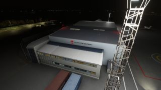 A Lufttransport hangar stands illuminated at night beside a lattice tower, with shipping containers in front and a parked aircraft nearby.