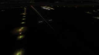 White lights outline a runway at night, with illuminated airport buildings nearby and scattered lights beneath distant mountain silhouettes.