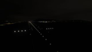 White runway lights extend toward illuminated airport buildings at night, with blue taxiway lights and dark hills beyond.