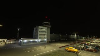 An airport control tower stands beside a terminal at night, overlooking illuminated roads and parked cars, including a yellow pickup truck.