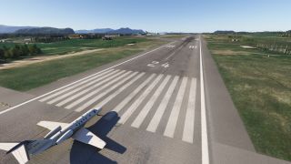A white business jet sits at runway 24’s threshold, with grassy verges, airport buildings, and distant mountains under clear skies.