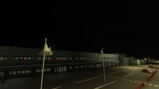 An airport terminal stands at night beside flagpoles, parked cars, pedestrian crossings, and a streetlit access road with red barriers.