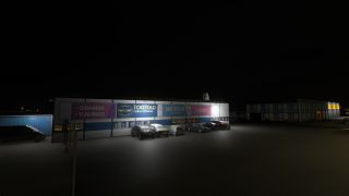 Parked cars line an illuminated Togstad Cargo Services building at night, with destination banners above its windows and fencing in the foreground.
