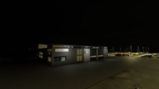 A gray building with illuminated windows and two garage doors stands beside a lamplit parking lot filled with vehicles at night.