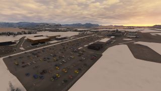 A snow-covered airport has large car parks, terminal buildings, and parked airliners, with water and mountains beneath an orange, cloud-filled sky.