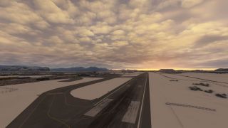 A runway and adjoining taxiways cross snow-covered airport grounds, with terminal buildings, distant mountains, and golden clouds along the horizon.