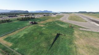 An airport runway borders grassy terrain, with connecting taxiways, parked airliners, terminal buildings, and distant mountains beneath a clear sky.