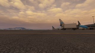 A KLM jet sits beside a terminal and service truck, with snow-covered mountains beneath an orange, cloud-filled sky.