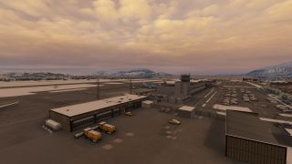 An airport terminal and control tower overlook parked jets, service vehicles, and a crowded car park beneath snowy mountains and orange clouds.