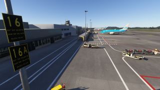 A KLM jet sits beside an airport terminal, with baggage carts on the apron and stand signs 16 and 16A in the foreground.