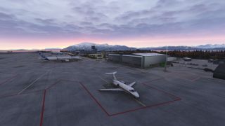 A white business jet sits on a marked apron near airliners and hangars, with snow-covered mountains beneath a pink, cloudy sky.