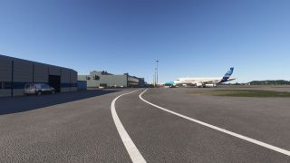 An Airbus A320 stands on the apron beside airport buildings and service vehicles, with curved white road markings in the foreground.