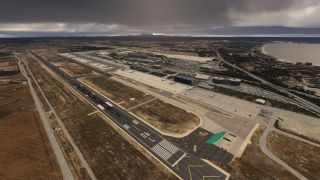 A coastal airport has a long marked runway, parked aircraft on broad aprons, and terminal buildings beneath dark clouds.