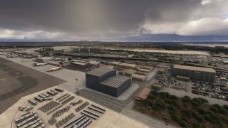 An airport apron contains parked airliners, hangars, rows of baggage carts and buses, with crowded car parks beneath dark clouds.