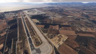 A coastal airport features runway 24L, parallel taxiways, and terminal aprons, with surrounding farmland and mountains beyond the shoreline.