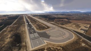 Runway 24L extends beside a parallel taxiway, surrounded by dry fields, with distant mountains and coastline beneath cloudy skies.