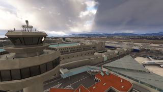 An airport control tower overlooks terminal buildings, parked airliners, and parallel taxiways, with distant mountains beneath heavy clouds.