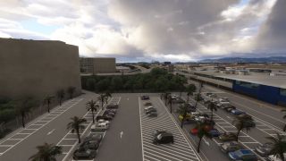 A palm-lined airport parking lot contains rows of cars beside terminal buildings, with aircraft and distant hills beneath heavy clouds.