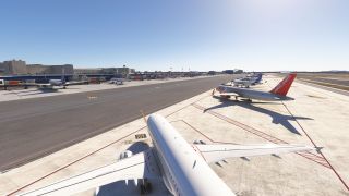 Several airliners occupy marked apron stands, including a red-tailed Jet2 aircraft, while terminal buildings and jet bridges line the opposite side.