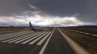 A twin-engine passenger jet sits on runway threshold markings beneath dark clouds, with airport buildings and distant hills along the horizon.