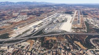 An airport has two runways, extensive terminal aprons, and highway access, surrounded by farmland and distant mountains.