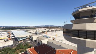 An airport control tower overlooks terminal buildings, jet bridges, and parked airliners, with runways and distant hills beneath clear blue skies.