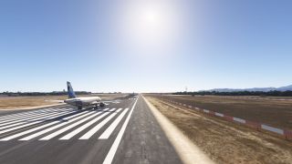 A twin-engine passenger jet sits on runway threshold markings, with dry grass, red-and-white barriers, and distant mountains under clear skies.