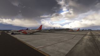 A Jet2 airliner stands on the apron beside ground vehicles, with other parked jets, terminal buildings, and dark clouds behind it.
