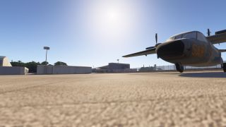 A gray twin-propeller aircraft with yellow SAR lettering sits on the apron near hangars, fencing, and floodlight poles.