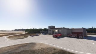 Red airport fire trucks sit outside a fire station beside curving service roads, with terminal buildings visible in the distance.