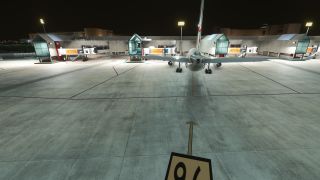 A rear view shows a twin-engine airliner parked on a floodlit apron at night, facing terminal buildings and jet bridges.