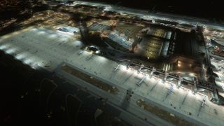 An aerial nighttime view shows aircraft parked beside illuminated terminal gates, broad apron areas, taxiways, and surrounding access roads.
