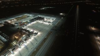 An elevated nighttime view shows floodlit airport terminals, parked airliners at jet bridges, and an adjacent runway outlined by lights.