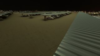 Rows of cars sit beneath long metal parking canopies at night, with distant lights visible beyond the concrete lot.