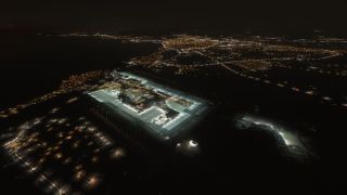 An aerial nighttime view shows illuminated airport terminals and aprons, green taxiway lights, and city lights tracing the coastline.
