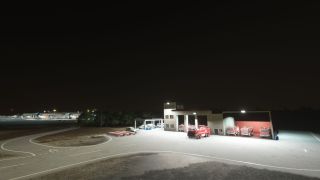 An illuminated airport fire station houses red fire engines beside curved service roads, with terminal buildings visible in the night background.