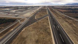 A coastal airport runway extends alongside taxiways and terminal aprons with parked airliners, while mountains rise beyond the shoreline.
