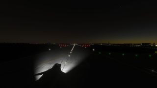 An aircraft sits on illuminated pavement at night, with white runway lights ahead and green taxiway lights beside distant airport buildings.