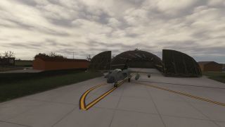 A gray military jet sits on yellow taxiway markings outside an open arched hangar beneath an overcast sky.