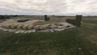 An airfield compound contains green containers, parked vehicles, hangars, and large curved ventilation ducts beneath an overcast sky.