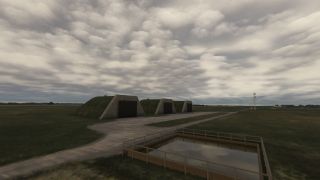 Three grass-covered concrete shelters stand beside paved paths, with a railed rectangular water basin in the foreground beneath an overcast sky.