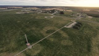 An airfield contains grass-covered shelters, hangars, circular paved areas, and a lattice tower amid open fields beneath a cloudy sky.