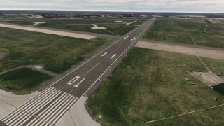 Runway 10 extends across a grassy airfield, with white threshold markings, intersecting taxiways, and distant buildings beneath an overcast sky.