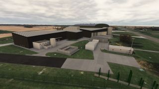 Large dark hangars stand beside storage tanks, utility buildings, and paved service areas, with taxiways and farmland beyond.