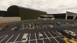 Cars occupy a marked parking lot beside a security fence, with a large green hangar and angular buildings beneath overcast skies.