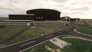 A large curved-roof building stands beside branching access roads, grassy verges, and a crowded parking lot under cloudy skies.